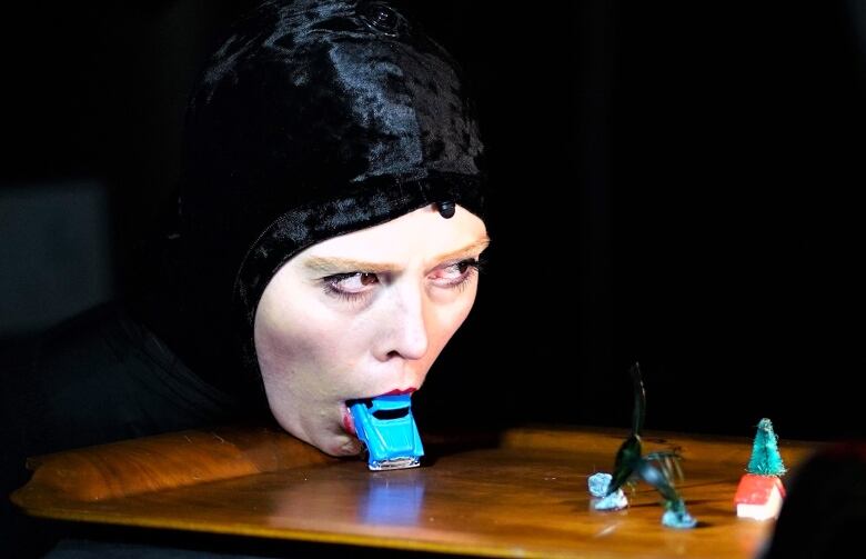 A closeup of a woman wearing a black hood with only her face exposed, resting her chin on a table. She has a small blue toy car in her mouth, and other miniatures sit on the table.