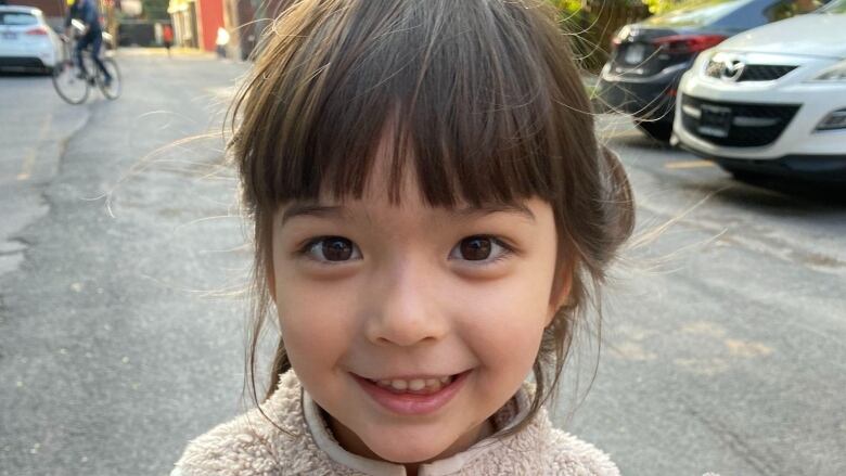 An adorable 4 year old girl smiles for the camera on a street. 