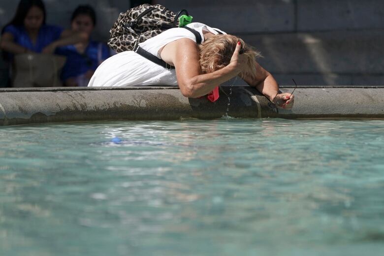 A woman ducks the top of her head into a pool of water at the base of a fountain. She is wearing a summer dress and is holding her glasses in her hand. 