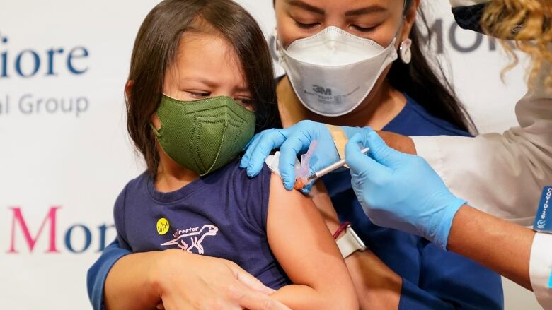 A woman wearing a face mask holds her young child, also wearing a face mask, while a nurse gives the child a shot in the arm with a needle.