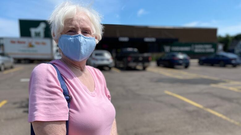 Woman with white hair and wearing face mask stands in a parking lot outside a brown brick building. 