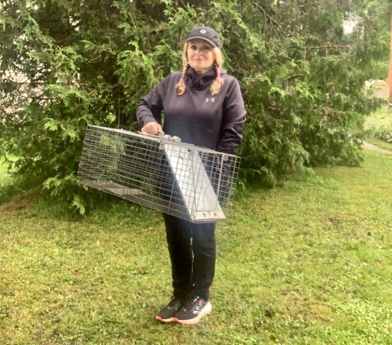 A woman stands in front of a tree holding a large wire-made cage. She wears a ball cap and a black jacket.