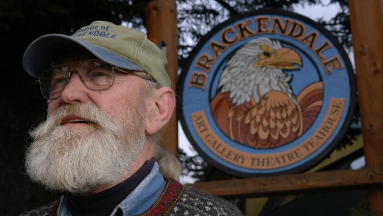 An old man with a white beard poses in front of an eagle symbol.