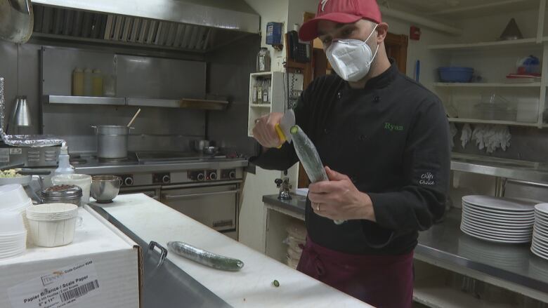 Man wearing face mask in commercial kitchen unwraps a cucumber with a knife.