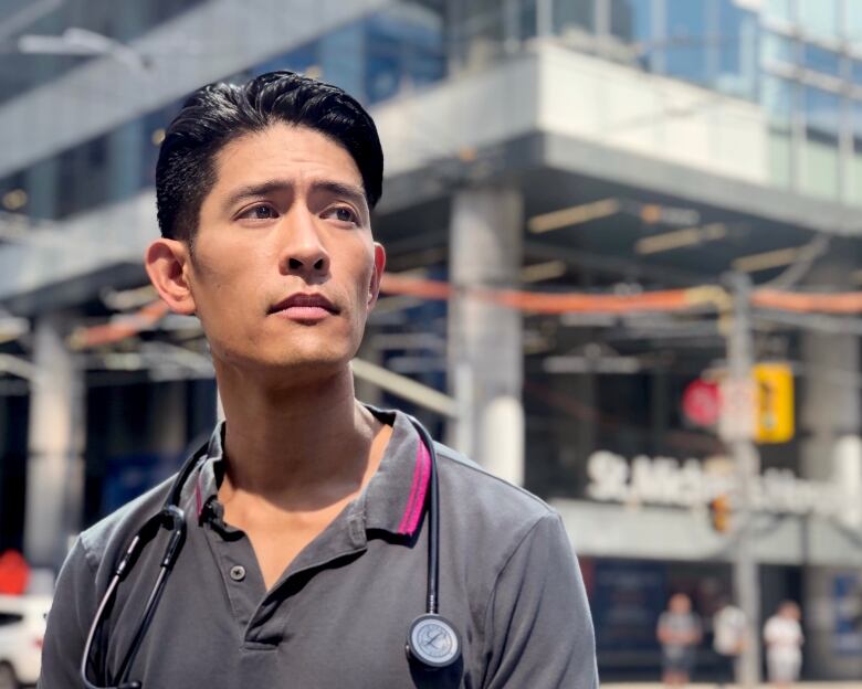 Man with short black hair and wearing grey collared shirt with stethoscope around neck stands in front of a hospital, looks into the distance.