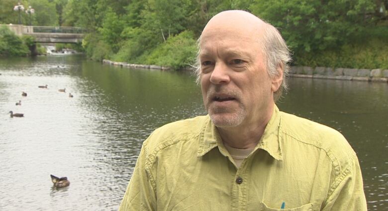 A man in a yellow button up shirt stands in front of a pond with ducks. 