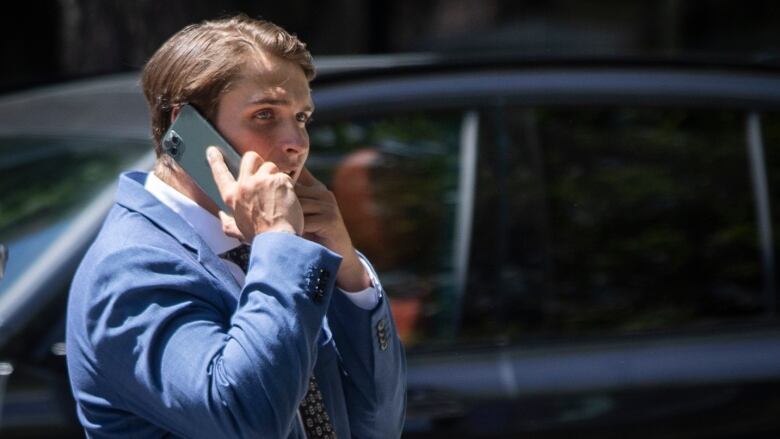 Jake Virtanen holds up a mobile phone to his ear. He is a white man with slicked-back brown hair, wearing a blue suit.