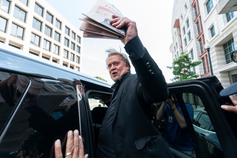 A man, with a folded newspaper in his hand, waves as he steps into a vehicle. 