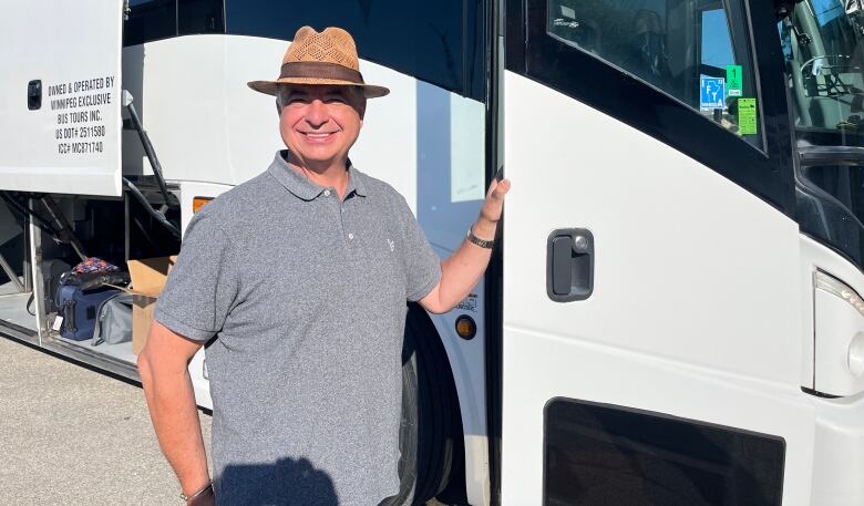 A man wearing a straw fedora hat poses, smiling, next to a white charter bus.