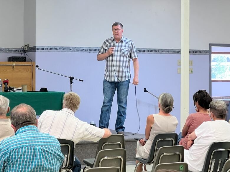 A man in blue jeans and a checkered shirt stands on a stage holding a microphone, speaking to a group of seated people.