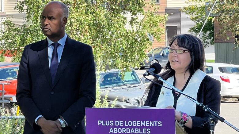 A woman speaks at a podium while a man stands beside her, listening.