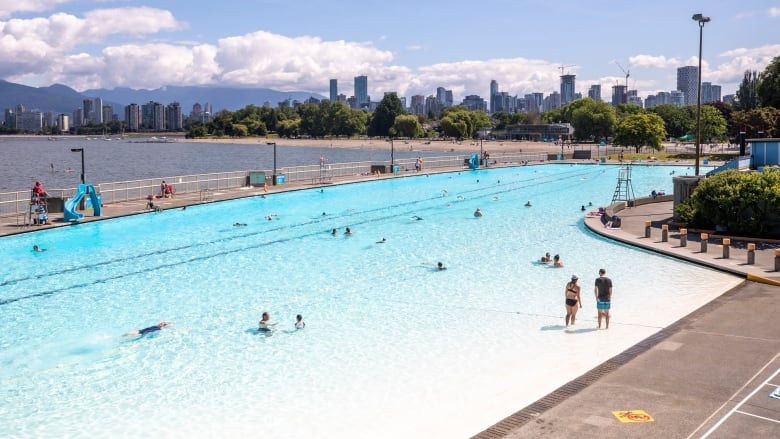 People swim and play in a large outdoor pool on an oceanfront beach with a cityscape in the background.