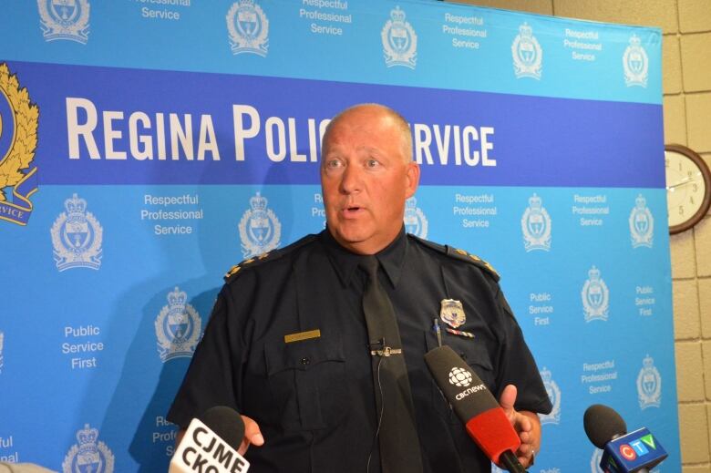 Regina Police Service Chief Evan Bray stands in front of a blue background emblazoned with the service's logo and motto. 