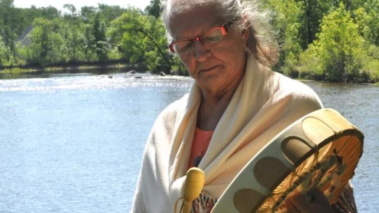 A woman holds a hand drum in front of a river.