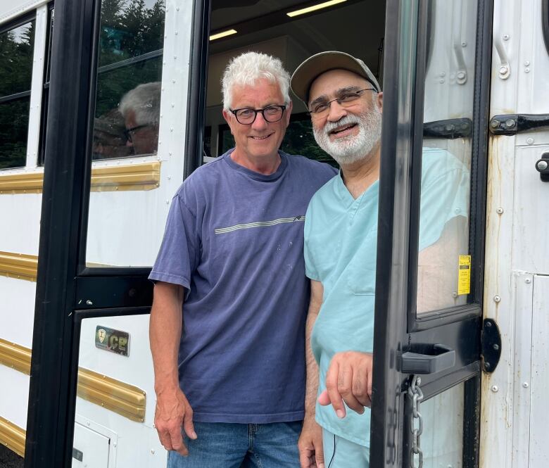 Two middle-aged men stand in the doorway of a bus, slightly leaning out and smiling into the camera.