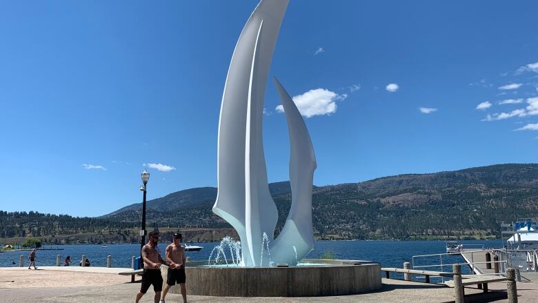 Two men walk past the large white Kelowna Sails sculpture next to Okanagan Lake on a sunny day.