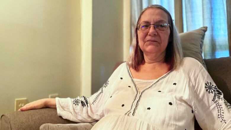 A woman in glasses and a white flowy dress sits on a couch looking into the camera.