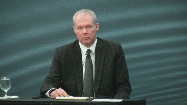 A man in a suit sits at a desk with a notebook and a glass of water in front of him.