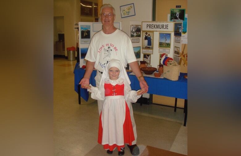 A young girl in a bright red dress with a white head covering stands in front of an older man, holding his hands.