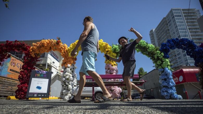 Two men smile and one flashes the rock on sign as they walk past a rainbow flower arrangement for Yaletown Pride in Vancouver, B.C.