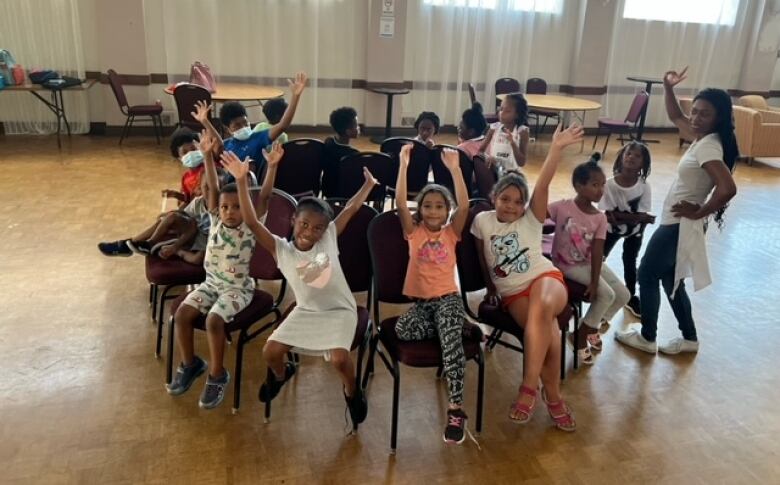 A group of children sit in a circle and wave at the camera.