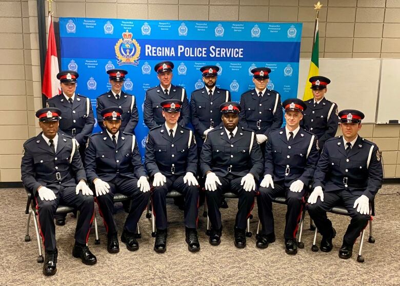 A group of 12 new Regina Police Service recruits sit and stand in front of a backdrop with the Service's logo on it and the Canadian and Saskatchewan flags. All wear their dress uniforms, hats, and white gloves. Nine are male and three female.