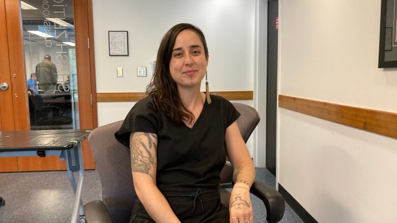 A woman with brown hair is seen sitting in a small classroom. She is wearing a black shirt and pants and has many visible tattoos on her arms, hands and face.