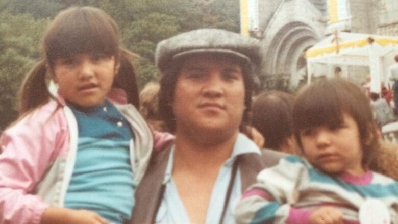 Picture of an Indigenous man holding his two young daughters in front of a church.