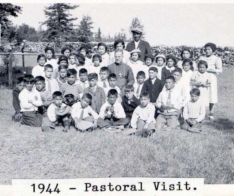 Black and white picture of a class of Indigenous children in residential school. A priest is in the middle of the group of children. There is sticker saying 