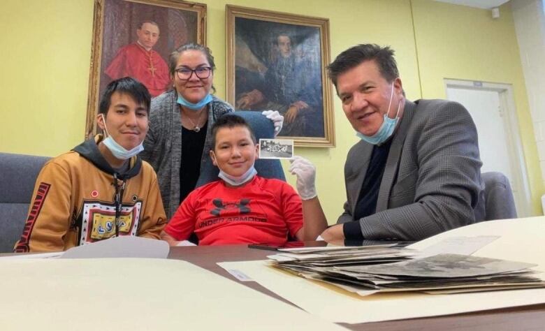 Picture of two children, their mother and their grandfather sitting at a table that has folders on it. One of the children is holding up a black and white photo. 