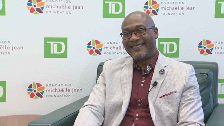 A Black man sitting in a green chair wearing glasses and a cream blazer