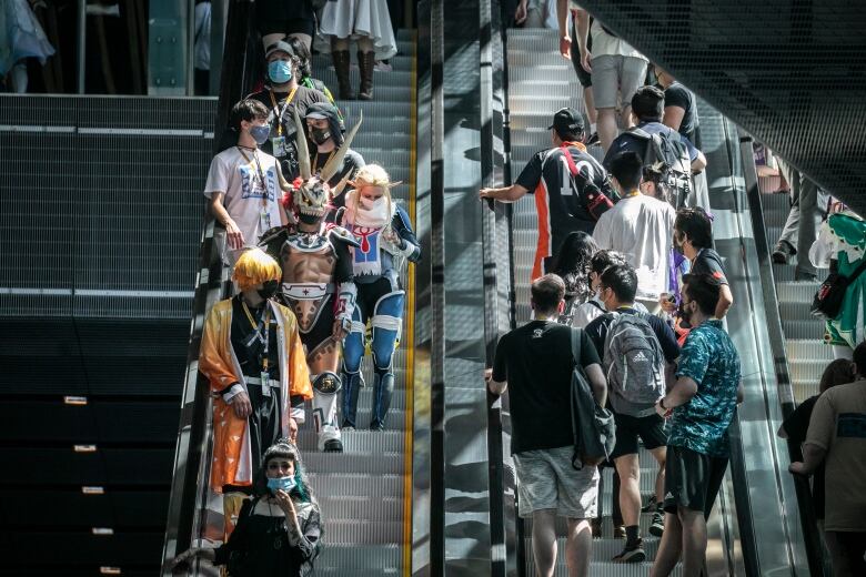 Two groups of people pass each other on an escalator. On the left side, people are pictured dressed up in amazing cosplays of Japanese characters.