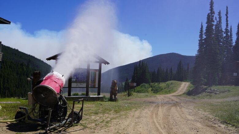 A snowblower blows mist over a small wooden structure, with a model bear next to it.