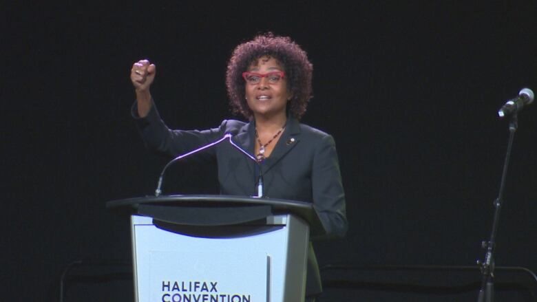 A Black woman with dark, curly hair is wearing a black blazer. She is speaking at a podium with a mic