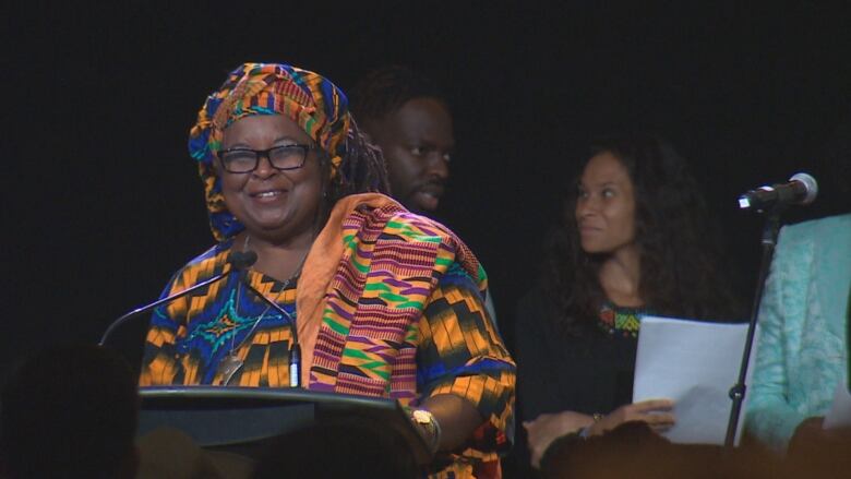 A Black woman with glasses wearing a colourful headdress and dress