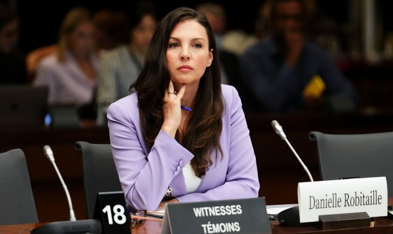 A woman in a suit sits at a long table, in front of a microphone.