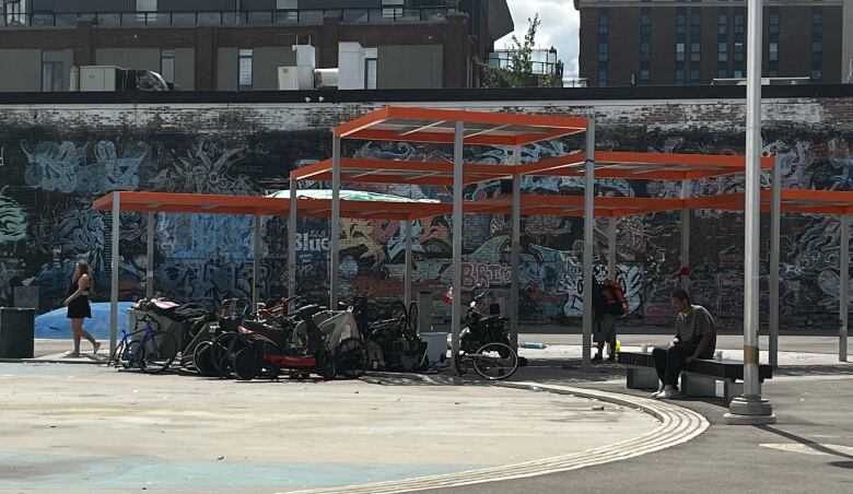 An orange shade overhang provides shade to a collection of bicycles. Behind, there is a wall covered in artistic grafitti. 