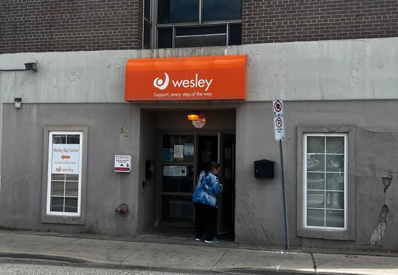 A beige building with a woman in blue scrubs entering. Above, an orange sign that says 'Wesley: Support every step of the way'.