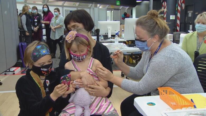 A child holding a stuffed toy, wearing a pink dress, gets a shot in the arm from a nurse.