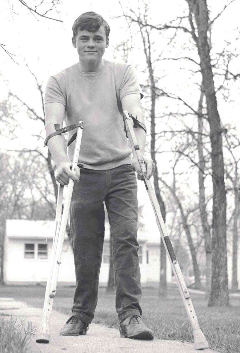 A teenaged boy stands on a sidewalk, leaning on a pair of crutches.