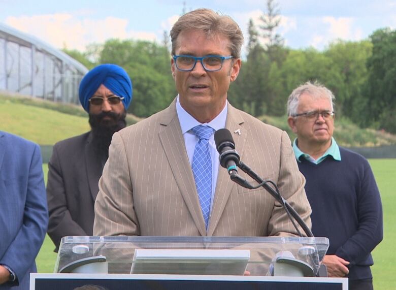 Mayoral candidate Kevin Klein speaks at a podium in Assiniboine Park, with two male supporters standing behind him.