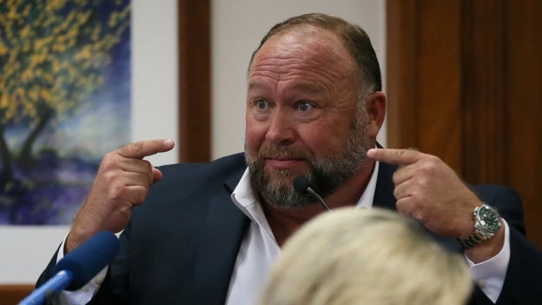 A man in a suit is shown inside a courtroom, in front of a mic, pointing at himself.