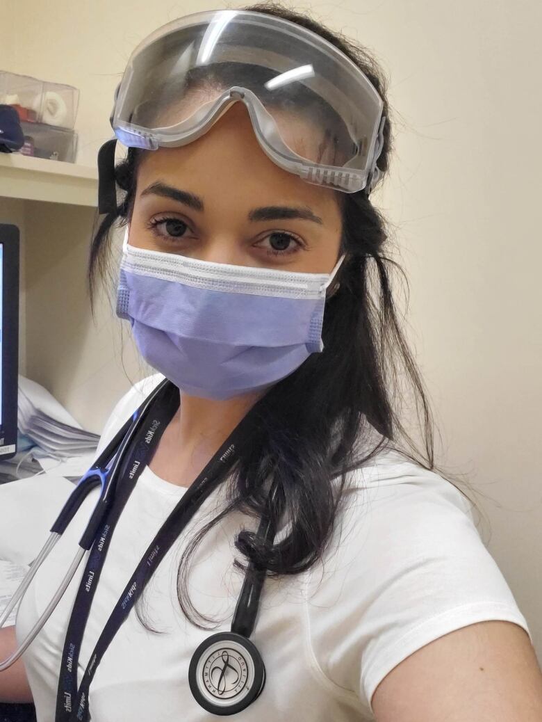 A healthcare worker wearing a scope, scrubs and a mask looks at the camera.