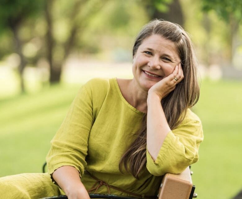 A portrait shot shows a woman sitting on a bench in a park. She's wearing a green dress, turned sideways to look at the camera, with her arm on the back of the bench and her head resting in her hand.