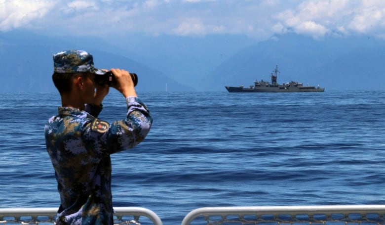 A soldier looks through binoculars at a ship across the water.