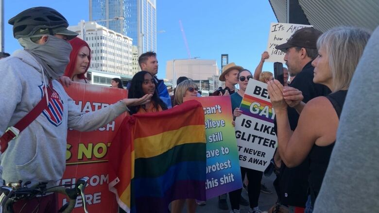 People holding a Pride flag and signs with messages of support are facing other people. One person is recording on their smartphone.