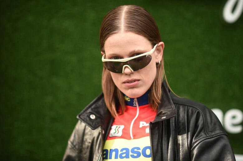 Kelly McCormack poses for the camera. She is wearing a NASCAR jersey under a black leather jacket, with large rectangular glasses.