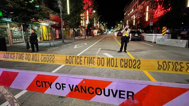 Two police officers stand behind crime tape on a closed-off street with hoardings and neon signs visible behind them.