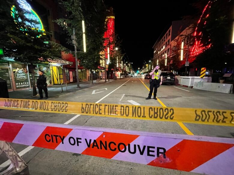 Two police officers stand behind crime tape on a closed-off street with hoardings and neon signs visible behind them.