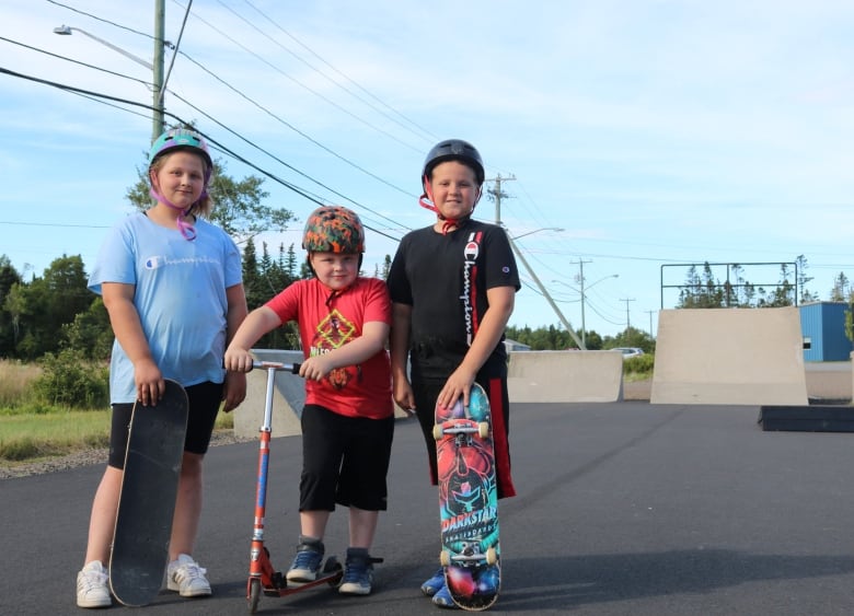 Three children wearing helmets and colourful T-shirts and shorts stand on a playground. Two are holding skateboards while one holds a scooter. 